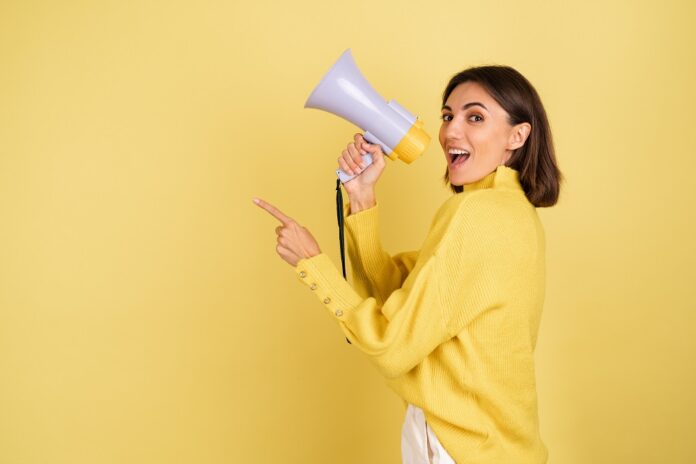 Young woman in yellow warm sweater with megaphone speaker screaming to the left point index finger happy excited